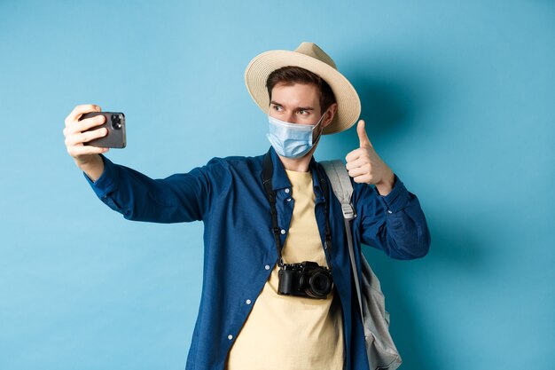 Covid-19, conceito de pandemia e viagens. feliz turista masculino tirando fotos de férias, faça selfie com o polegar para cima, recomendando o hotel, de pé sobre fundo azul.