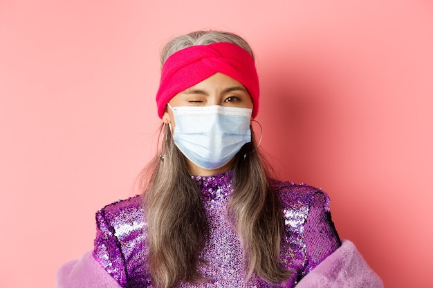 Covid-19, conceito de vírus e distanciamento social. Atrevida mulher asiática sênior na máscara médica e vestido glitter piscando, vestindo roupa de festa, fundo rosa.