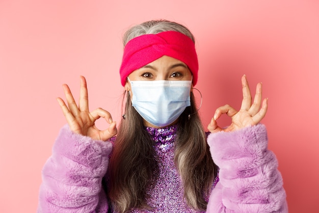 Covid-19, conceito de vírus e moda. Close-up de elegante mulher asiática sênior na máscara facial, mostrando sinais de ok e olhando um fundo rosa, feliz.