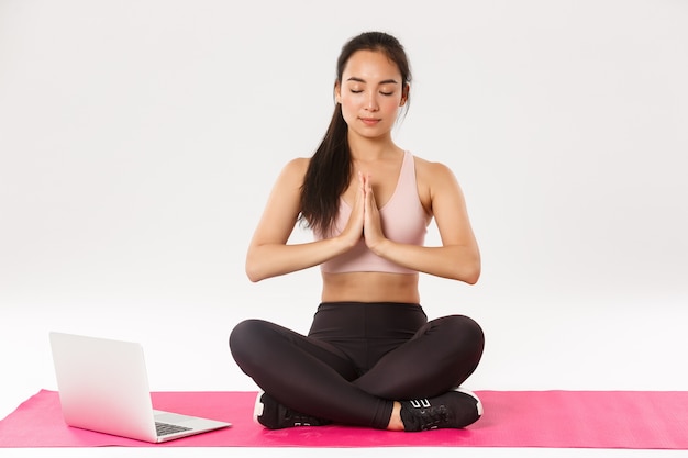 Covid-19 y concepto de bienestar. Retrato de una chica asiática sonriente relajada que se queda en casa, se pone en cuarentena y practica yoga, sentada en una alfombra de goma en posición de loto, repite después del video en la computadora portátil.