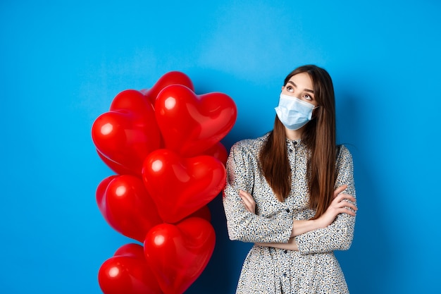 Covid-19, concepto de cuarentena y salud. Hermosa niña soñadora en mascarilla y vestido, mirando pensativo en la esquina superior izquierda, de pie cerca de globos del día de San Valentín, fondo azul