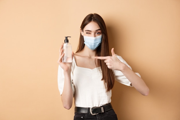 Covid-19 y concepto de medidas preventivas. Niña sonriente guiñando un ojo en máscara médica, apuntando a la botella de desinfectante de manos, mostrando un buen producto, fondo beige.