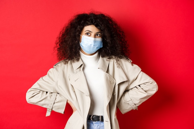 Covid-19, concepto de pandemia y cuarentena. Mujer elegante y entusiasta en máscara médica y gabardina, sonriendo y mirando a la esquina superior izquierda, fondo rojo.