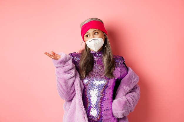 Covid-19, concepto de pandemia y moda. Elegante mujer mayor asiática en respirador y vestido de fiesta mirando escéptico, apuntando con la mano hacia la izquierda, de pie sobre fondo rosa