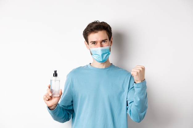 Covid-19, concepto de salud y cuarentena. Hombre alegre en mascarilla celebrando, mostrando la bomba de puño y la botella con desinfectante de manos, luchando contra los gérmenes, fondo blanco.