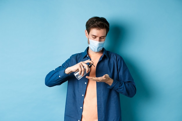Covid-19, concepto de salud y pandemia. Chico joven en máscara médica limpiando las manos con desinfectante, aplicar antiséptico en la palma, de pie sobre fondo azul.