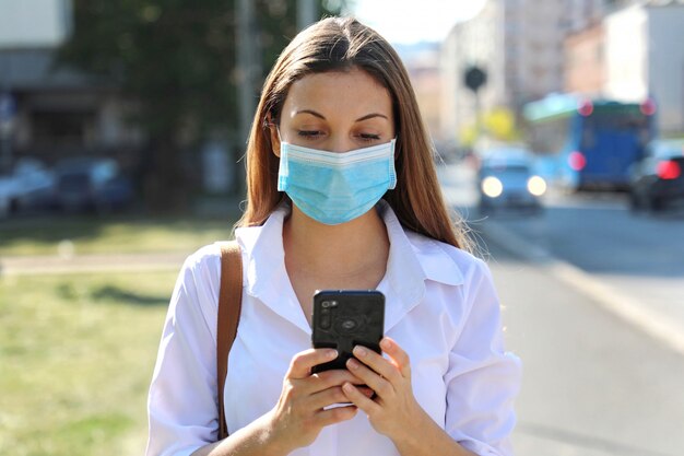 COVID-19 Coronavirus pandémico Mujer joven con máscara quirúrgica usando la aplicación de teléfono inteligente en City Street