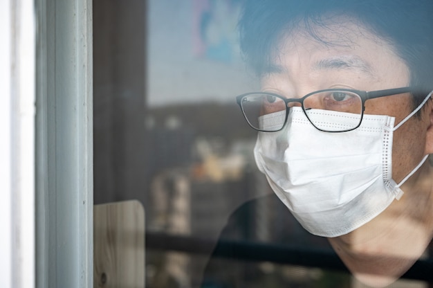 COVID-19 Coronavirus-Selbstisolationskonzept. Ein asiatischer Mann mit einer Gesichtsmaske und einem Fenster, das während seiner Selbstisolation zu Hause aus dem Fenster schaut.