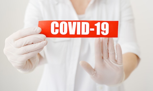 COVID-19 Coronavirus, sinal de aviso vermelho com texto COVID-19 nas mãos do médico de jaleco branco, luvas, máscara médica do rosto. Coronavírus trimestral Epidemia de gripe epidêmica. COVID19 Conceito médico de cuidados de saúde