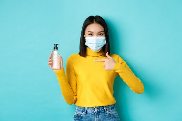 Covid-19, distanciamento social e conceito de pandemia. Mulher asiática alegre mostrando desinfetante para as mãos, recomendo o uso de anti-séptico de coronavírus, usando máscara médica.