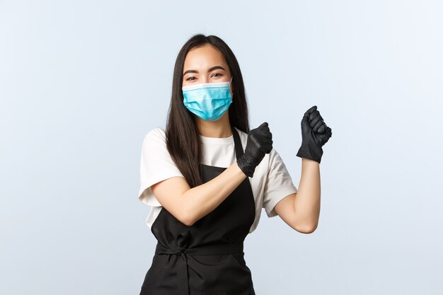 Covid-19, distanciamiento social, pequeño negocio de cafetería y concepto de prevención de virus. Feliz sonriente barista asiática, empleada de café bailando anticipando visitantes, use mascarilla médica y guantes