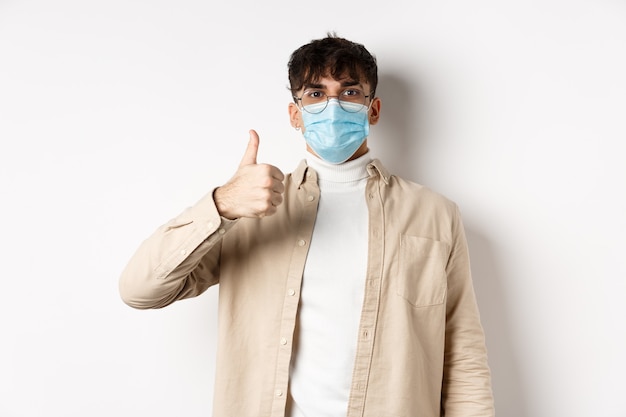 Covid-19, Gesundheits- und Real-People-Konzept. Zufriedener Mann in steriler Gesichtsmaske und Brille, Daumen hoch in Zustimmung, positives Feedback geben, auf weißer Wand stehen.