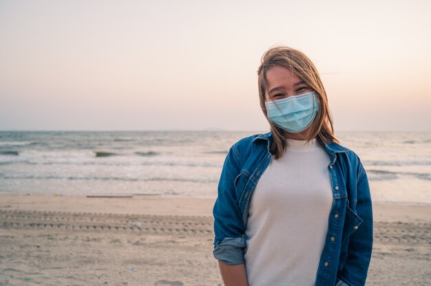 Covid-19-Konzept, asiatische junge Frau, die eine Hygieneschutzmaske über ihrem Gesicht im Freien mit Sonnenuntergang am Strand trägt