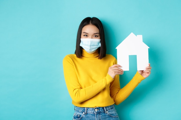 Covid-19, pandemia y concepto inmobiliario. Mujer asiática joven con ropa amarilla que muestra el recorte de la casa de papel, con máscara médica durante el coronavirus, de pie sobre fondo azul.