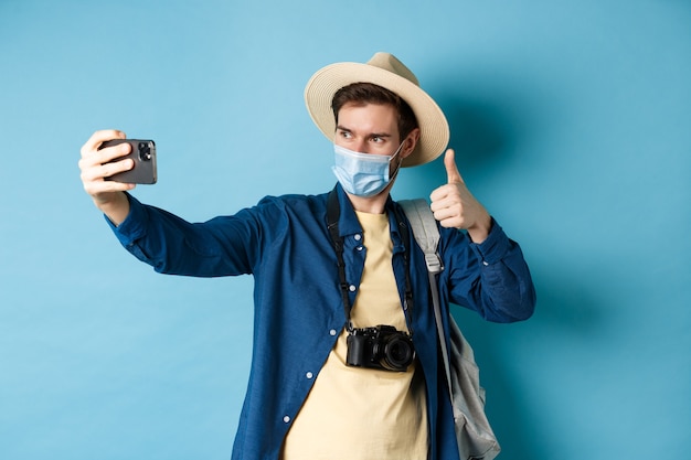Covid-19, pandemia y concepto de viaje. Turista masculino feliz tomando fotografías de vacaciones, hacer selfie con el pulgar hacia arriba, recomendando el hotel, de pie sobre fondo azul.