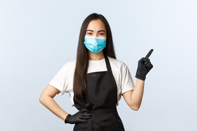 Covid-19 pandemia, distanciamento social, pequenas empresas e conceito de prevenção de vírus. Barista de café asiático sorridente, funcionário com máscara médica e luvas apontando o canto superior direito.