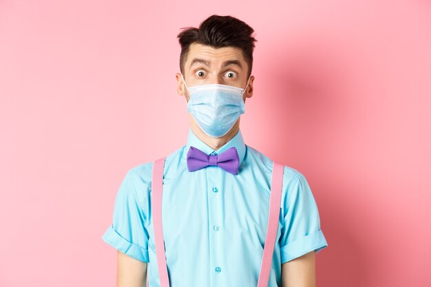Covid-19, pandemia e conceito de saúde. Imagem de cara parecendo surpreso com máscara médica e gravata borboleta, de pé sobre fundo rosa.
