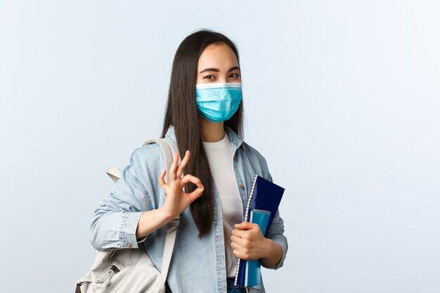 Covid-19-Pandemie, Bildung während des Coronavirus, zurück zum Schulkonzept. Zufriedene süße asiatische Studentin in medizinischer Maske, okaygeste als Studium an einer coolen Universität zeigen, Notizbücher halten.