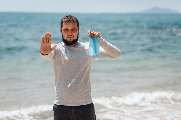 COVID-19 Pandemie Coronavirus junger Reisender am Strand mit Gesichtsmaske, der an seiner Hand "Stopp" zum Virus sagt, schützend für die Ausbreitung der Coronavirus-Krankheit 2020. SARS-CoV-2-Konzept