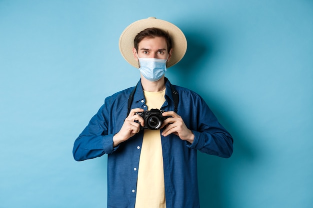 Covid-19, Pandemie und Reisekonzept. Fröhlicher Tourist im Sommerhut und in der medizinischen Maske, die Fotos machen, Kamera halten, auf blauem Hintergrund stehend.