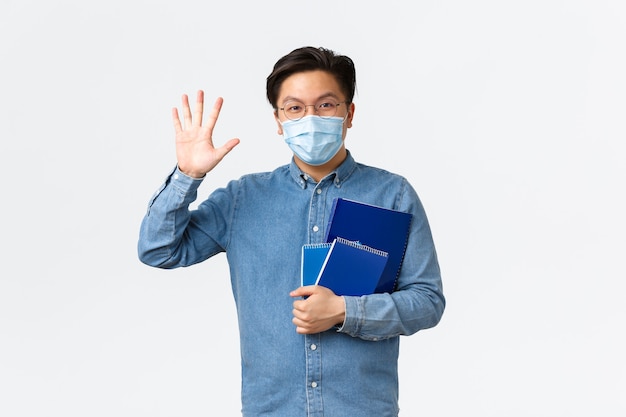 Covid-19, prevención de virus y distanciamiento social en el concepto universitario. Amistoso estudiante asiático sonriente diciendo hola, saludando con la mano en gesto de saludo, llevando cuadernos, usando máscara médica y gafas.