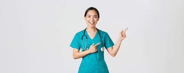 Covid-19, trabajadores de la salud, concepto de pandemia. Sonriente y alegre doctora asiática, terapeuta en matorrales señalando con el dedo la esquina superior derecha, invitando a un chequeo, recomienda vacunar.