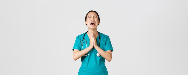 Covid-19, trabajadores de la salud, concepto de pandemia. Triste y cansada doctora asiática con exceso de trabajo, enfermera pidiendo ayuda, suplicando a Dios y llorando de estrés, de fondo blanco de pie