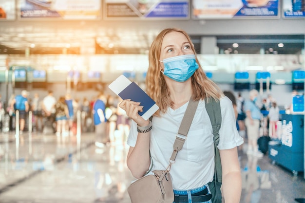 COVID-19 Turista de cabelo loiro feliz com uma mochila em uma máscara médica protetora com passaporte enquanto aguarda a verificação do passaporte do turista no aeroporto