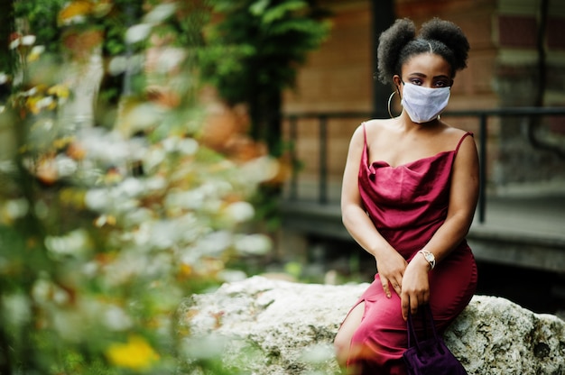 Covid-19, vírus infeccioso. Mulher africana com cabelo encaracolado, usa vestido de seda vermelho e máscara médica descartável, se preocupa com sua saúde e protege em situação perigosa.