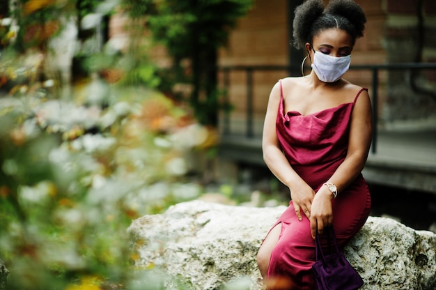 Covid-19, vírus infeccioso. Mulher africana com cabelo encaracolado, usa vestido de seda vermelho e máscara médica descartável, se preocupa com sua saúde e protege em situação perigosa.