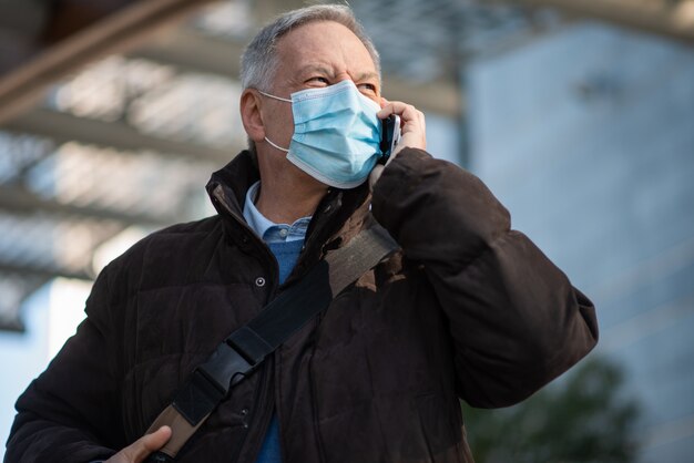 Covid Coronavirus-Konzept, maskierter älterer Geschäftsmann, der auf seinem Smartphone im Freien spricht