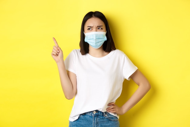 Covid-, Gesundheits- und Pandemiekonzept. Asiatische Frau in medizinischer Maske und weißem T-Shirt, die mit dem Finger auf das Logo in der oberen linken Ecke zeigt, Werbung zeigt, gelber Hintergrund