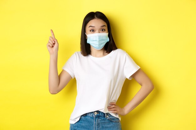 Covid-, Gesundheits- und Pandemiekonzept. Asiatisches weibliches Modell in medizinischer Maske und weißem T-Shirt, das mit dem Finger auf das Logo in der oberen linken Ecke zeigt, Werbung zeigt, gelber Hintergrund