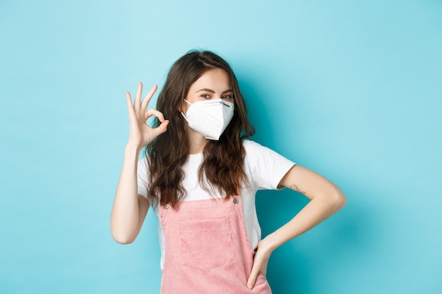 Covid, salud y concepto de pandemia. Muy bien. Joven mujer solidaria vistiendo un respirador médico y mostrando un signo de aprobación aceptable, alaban el uso de máscaras faciales en público, fondo azul