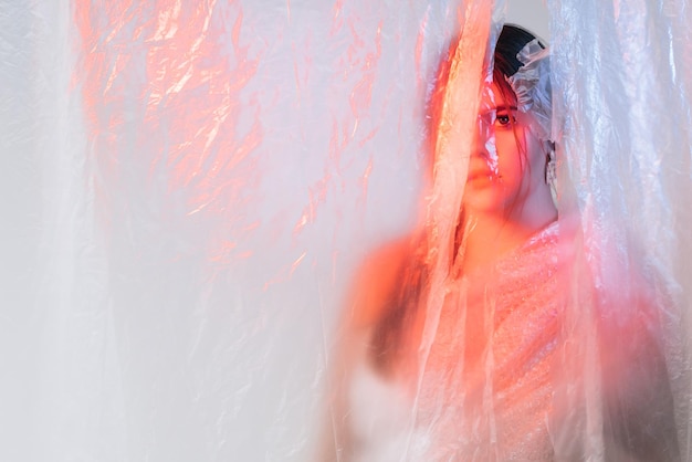 Covid19 autoaislamiento Riesgo de infección Soledad en cuarentena Retrato artístico de una mujer perturbada escondida detrás de una película transparente de polietileno arrugado en una luz de neón azul rosa iridiscente en un espacio vacío
