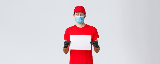 Covid19 Selbstquarantäne-Online-Shopping- und Versandkonzept Aufgeregter Kurier in roten Uniformhandschuhen und Gesichtsmaske mit leerem Blatt Papier und starrer überraschter Kamera mit Firmeninformationen oder Banner