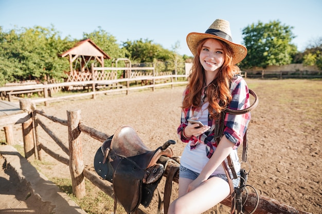 Foto cowgirl bonita sorridente em pé e usando o celular no rancho