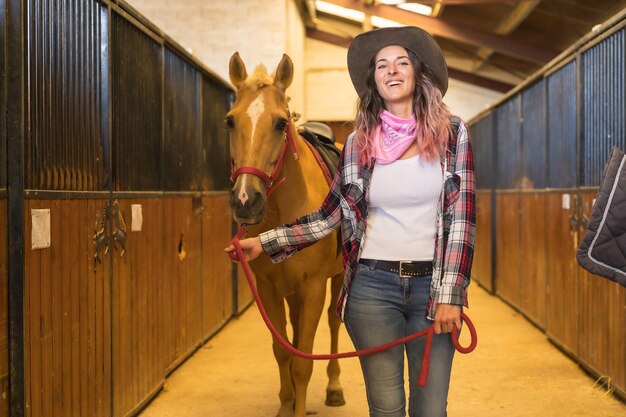 Cowgirl de cabelo rosa caucasiano passeando com um cavalo em um estábulo, chapéus de cowboy americanos, camisa xadrez rosa e jeans