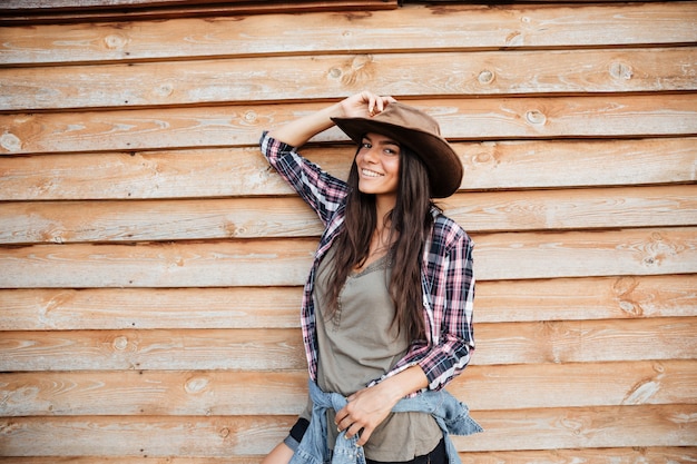 Cowgirl feliz e atraente jovem com camisa xadrez e chapéu em pé e sorrindo sobre um fundo de madeira.