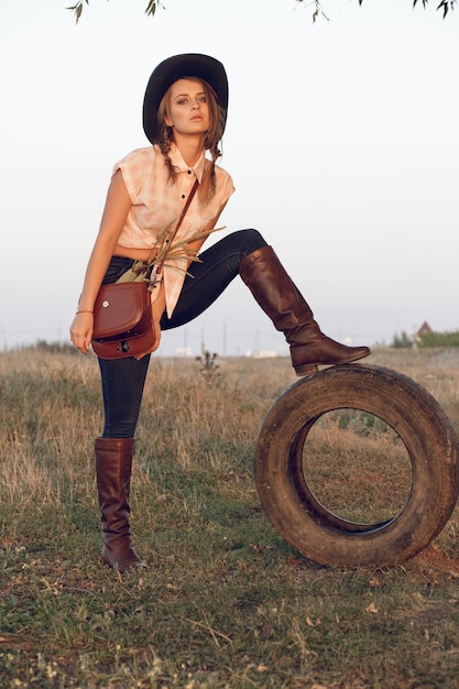 Foto cowgirl garota com camisa de botas jeans e com uma bolsa em uma caixa com uma roda perto de uma árvore