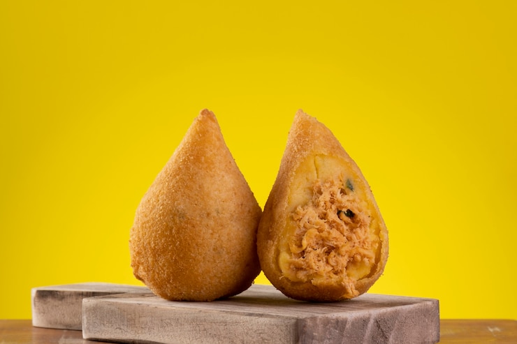 Foto coxinha frita em tábua de madeira com fundo amarelo.