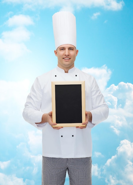 Foto cozinha, profissão, propaganda e conceito de pessoas - cozinheiro chef masculino feliz mostrando e segurando a placa de menu em branco sobre o céu azul com fundo de nuvens