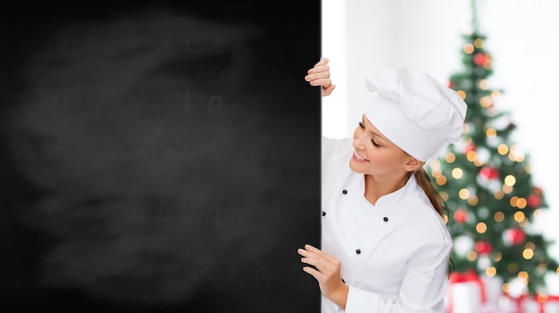 cozinha, propaganda, feriados e conceito de pessoas - sorridente chef feminina, cozinheira ou confeiteira com lousa em branco sobre a sala de estar e o fundo da árvore de natal