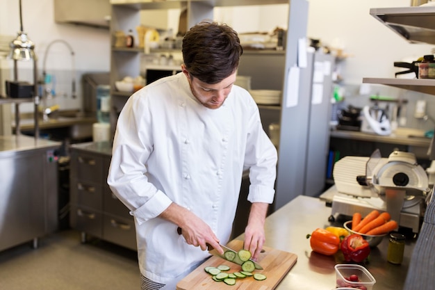 cozinhar alimentos, profissão e conceito de pessoas - cozinheiro chef masculino feliz com faca cortando pepino na tábua na cozinha do restaurante