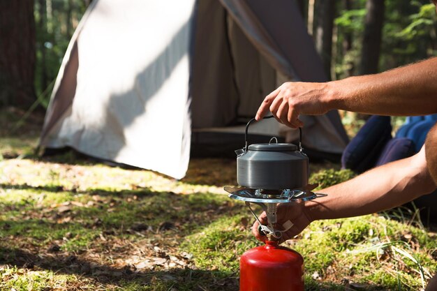 Cozinhar aquecendo uma chaleira turística em um queimador de gás portátil com um cilindro de gás vermelho acampar um homem cozinha o café da manhã ao ar livre atividades ao ar livre de verão