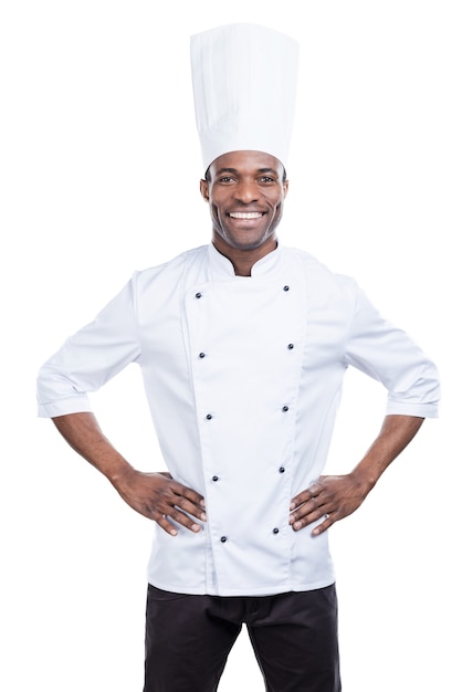 Cozinhar é minha vida! Jovem chef africano confiante de uniforme branco, segurando as mãos na cintura e sorrindo