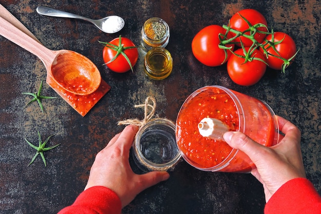 Foto cozinhar molho de tomate caseiro. tomate e especiarias. mãos femininas em mangas compridas vermelhas despeje o molho de tomate preparado na hora de um copo de liquidificador em uma jarra de vidro.