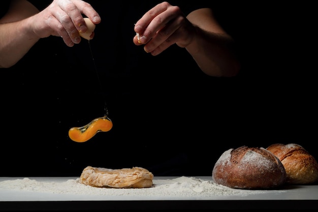 Cozinhar o pão. As mãos do chef colocam um ovo de galinha que voa para a mesa branca com a farinha e a massa. Chef polvilha pó de farinha branca com a mão de um homem em um fundo preto. Espaço para texto.