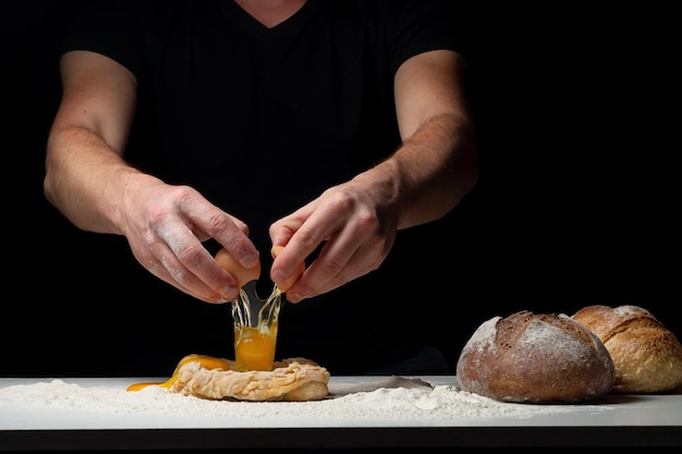 Cozinhar o pão. as mãos do chef colocam um ovo de galinha que voa para a mesa branca com a farinha e a massa. chef polvilha pó de farinha branca com a mão de um homem em um fundo preto. espaço para texto.