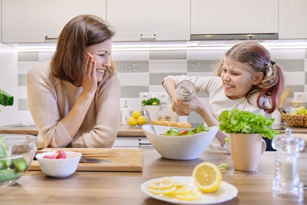 Cozinhar refeição saudável em casa por família. menina sais salada recém cozida, mãe olha para cima e se alegra